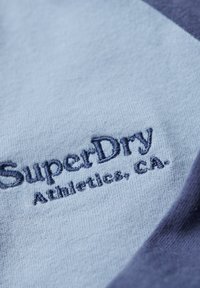 Hellblauer Stoff mit dunkelblau besticktem Text "SuperDry Adventure, CA." und einem dunkelblauen Rand, der auf der rechten Seite sichtbar ist.