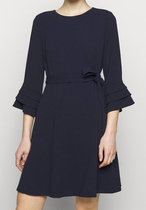 Femme portant une robe bleu marine longueur genou avec manches cloche à volants 3/4 et ceinture nouée à la taille, debout devant un fond uni.