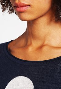 Pull bleu marine avec un col large, présentant un grand motif circulaire blanc ; fabriqué en maille douce avec une finition texturée.