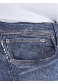 Tmavě modré džíny z denimu zobrazují detailní pohled na pas s měděnými nýtky a kontrastním šitím podél kapes a švů.