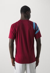 Bordeauxfarbenes Sportshirt mit kurzen Ärmeln und rundem Ausschnitt, das blaue Streifen auf der rechten Schulter aufweist und aus einem glatten, atmungsaktiven Stoff gefertigt ist.