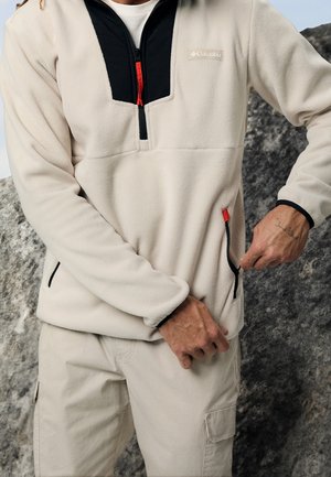 Personne portant un fleece Columbia beige et noir avec demi-fermeture éclair et poches avant, ajustant la fermeture éclair de la poche droite, debout devant un fond rocheux.