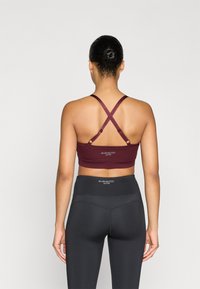 Bordová športová podprsenka s prekríženými ramienkami a detailom loga; v kombinácii s čiernymi legínami s vysokým pásom a jemným švovým vzorom.