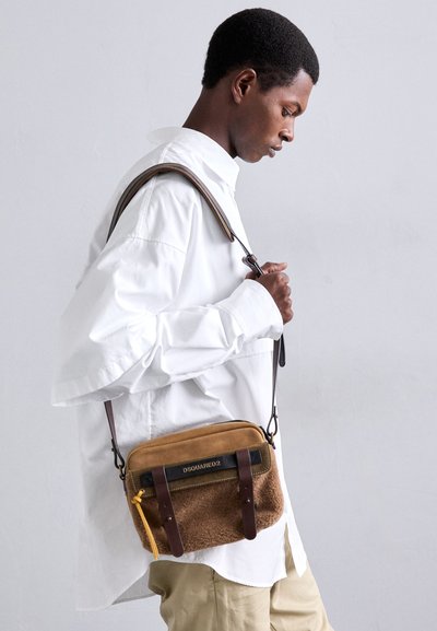 Geantă crossbody din material textil maro, cu o textură de blană de oaie, curele din piele și un fermoar auriu. Include un accent cu logo negru.