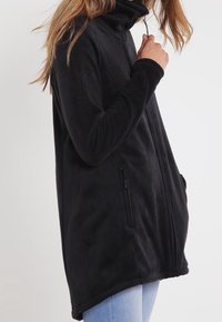 Mustat fleece-takit, joissa on korkea kaulus, vetoketjusulku, sivutaskut, pitkät hihat ja löysä, pitkänomainen muotoilu. Pehmeä rakenne, sileä pinta.