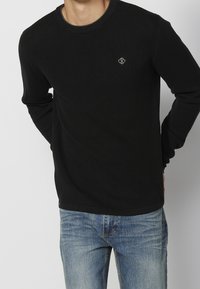 Černý žebrovaný svetr s dlouhými rukávy a kulatým výstřihem, s malým vyšitým logem na hrudi, v kombinaci se světle modrými džíny.