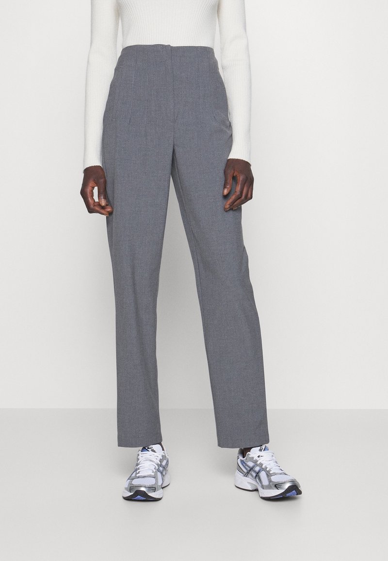 Grijze op maat gemaakte broeken met een hoge taille, riemlussen en plooien; gecombineerd met een witte geribde longsleeve en grijs/blauwe sportschoenen.