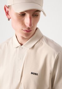 Sviestinė polo marškinėliai su tekstūruotu raštu ir trimis mygtukais, ant krūtinės esantis juodas "BOSS" logotipas. Pora su atitinkama kepure.