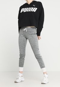 Cappuccio nero cropped con logo "PUMA" bianco, abbinato a pantaloni da jogging grigi a gamba stretta con strisce laterali e costine bianche alla caviglia.