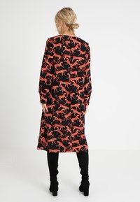 Langes schwarzes Kleid mit einem lebhaften roten Leopardenmuster, langen Ärmeln und einer entspannten Silhouette, kombiniert mit schwarzen kniehohen Stiefeln.