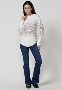 Pulover crem cu stropi multicolori, decolteu rotund, manșete cu rimă și margine curbată, asociat cu blugi evazați albaștri și pantofi negri.