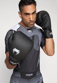 Luvas de boxe pretas com design acolchoado e logótipo branco, usadas por uma pessoa com roupa atlética texturizada em preto e cinza.