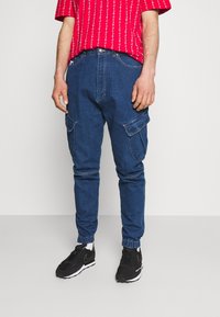 Pantalones cargo de mezclilla azul con puños estilo jogger, que presentan múltiples bolsillos y un corte relajado. Combinados con una camisa de manga corta de patrón rojo.