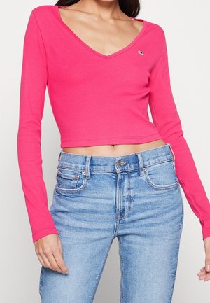 Kobieta ubrana w jasnoróżowy, długi crop top z dekoltem w kształcie litery V oraz jasnoniebieskie, wysokie spodnie jeansowe na tle białego, gładkiego tła.