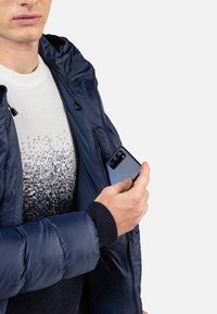 Giacca a vento blu navy con cappuccio, dotata di chiusura con zip e polsini a coste. Sotto indossa un maglione con gradiente bianco e blu navy. Smartphone in mano.