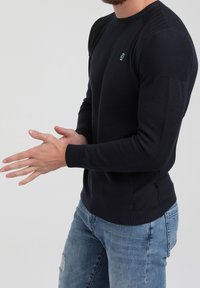 Pull en maille bleu marine avec tissu texturé, poignets côtelés et col rond. Comprend des fentes latérales et un petit logo sur la poitrine.