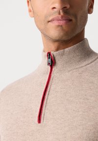 Beige sweater with a high collar, featuring a red zipper and contrasting stitching. The texture is smooth with a soft, knitted finish.