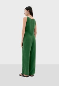 Un combinaisons en lin vert avec un design sans manches, des jambes larges et une fermeture éclair dans le dos. Il possède une texture lisse et un col modeste.