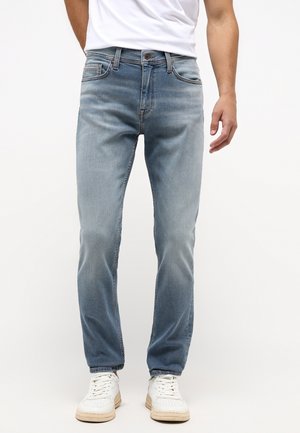 Mann trägt hellblaue Slim-Fit-Jeans, weißes T-Shirt und weiße Sneakers, steht vor einem schlichten weißen Hintergrund.