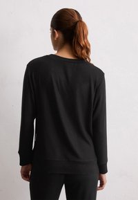 Donna con capelli castani legati in una coda di cavallo, che indossa un semplice maglione nero a maniche lunghe e pantaloni neri, di fronte a un muro bianco texturizzato.