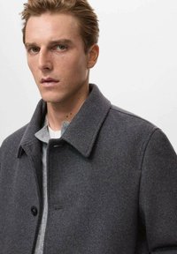 Abrigo de lana gris con un diseño recto, cuello ancho, cierre de botones en la parte delantera y una textura suave, superpuesto sobre una camisa gris clara.