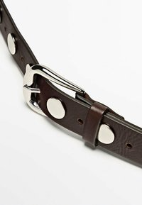 Bruine leren riem met een zilveren metalen gesp, voorzien van ronde metalen accenten langs de band. Gladde textuur met zichtbare stiksels.