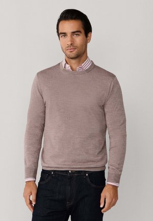 Suéter marrón claro hecho de un material de punto, con cuello redondo y mangas largas, usado sobre una camisa de cuello a rayas y jeans oscuros.