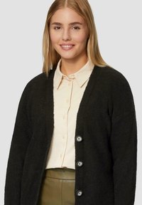 Schwarzer, flauschiger Cardigan mit strukturierten Oberflächen, V-Ausschnitt, drei großen Knöpfen und getragen über einem hellbeigen Hemd mit Knöpfen.