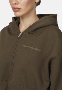 Felpa con cappuccio marrone con zip, caratterizzata da un tessuto testurizzato e la frase "GOODFORNOTHING" in un verde chiaro.