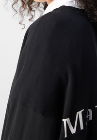 Czarna bluza z białym napisem; miękka tkanina, luźny krój, widoczne detale przeszycia, okrągły dekolt i teksturowane rękawy.