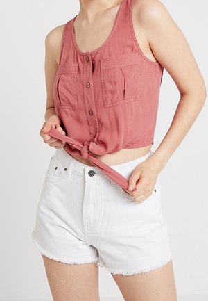 Vrouw die de onderkant van een mouwloos roze hemd met knopen vastknoopt, draagt witte denim shorts met hoge taille en rafelige zoom.