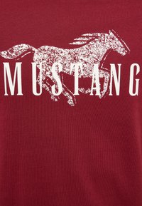 Silhouette blanche usée d'un cheval au galop derrière le mot « MUSTANG » en lettres majuscules sur un fond de tissu rouge.