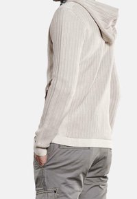 Light beige knit hoodie with ribbed texture, long sleeves, and a kangaroo pocket. Features a neutral cuff and hem. Paired with gray cargo pants.