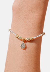 Bracelet en or et argent avec des perles blanches, présentant un charme en forme de goutte d'eau. Notable pour ses accents dorés texturés et ses éléments de design complexes.