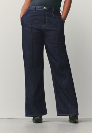 Person trägt dunkelblaue Jeans mit weitem Bein und vorderen Nähten, die Hände in den Taschen, dazu schwarze Stiefel und ein dunkles Oberteil.