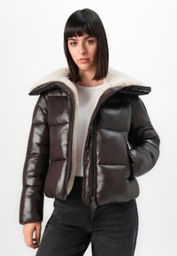Vrouw met stijlvol donker haar, gekleed in een gedeeltelijk opengeritste donkerbruine pufferjack met een witte fleece voering en zwarte broek, staande tegen een witte achtergrond.