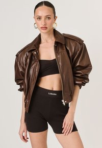 Barna bőrkabát párnázott vállakkal, zárt cipzárral, és bordázott fekete rövidnadrággal, logós derékrésszel. Sima textúra és modern szabás.