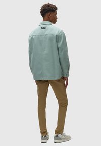 Mintgroene, button-up jas met een kraag, achteraan een logo tag, en lange mouwen. Gecombineerd met tan chinos en witte sneakers.