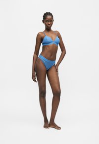 Grande femme aux cheveux tressés portant un soutien-gorge bleu texturé et une culotte taille haute assortie, debout pieds nus sur un fond gris uni.