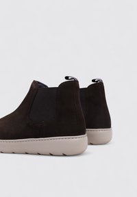 Bottines en daim marron avec des panneaux latéraux élastiqués et une semelle en caoutchouc beige. L'arrière est doté d'une languette avec un accent rouge et bleu.
