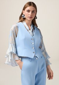 Gilet azzurro chiaro con sei bottoni e tasche laterali, abbinato a una blusa a strisce fluida con maniche a volants in morbidi colori pastello.