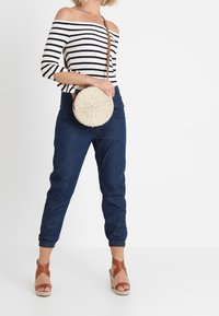 Žena v pruhovaném topu s odhalenými rameny, tmavě modrých sportovních kalhotách, světlehnědých klínových sandálech a s kulatou tkanou kabelkou přes rameno.