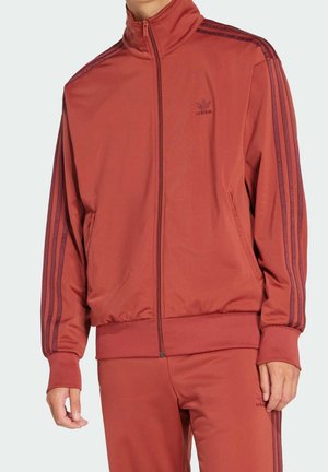 Persoon draagt een roestkleurig Adidas trainingspak met een ritsvest en bijpassende broek, beide met donkerrode strepen op de mouwen en benen.