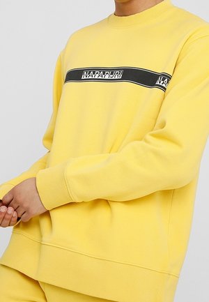 Person iført en gul sweatshirt med en sort vandret stribe og teksten "NAPAPIJRI" henover brystet, hænder løst sammenslået.