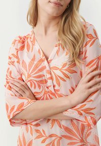 Ljusrosa blus med blommönster i nyanser av orange. Har V-urringning och trekvartsärmar. Tillverkad av mjukt, transparent tyg.