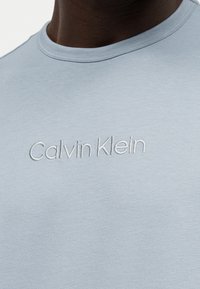 Κοντινό πλάνο ενός γαλάζιου T-shirt με στρογγυλή λαιμόκοψη και λευκό κεντημένο λογότυπο «Calvin Klein» στο στήθος.