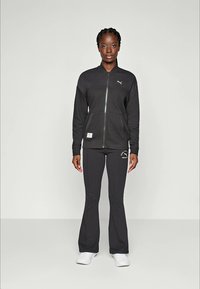 Svart zip-up jacka med PUMA-logotyp och två fickor, kombinerad med svarta utsvängda byxor med en logotyp vid vristen, bärs med vita sneakers.