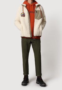 Krémová fleecová bunda se zelenými akcenty, červenou kapucí, olivově zelené kalhoty a černé boty. Má náprsní kapsu a kontrastní šití.