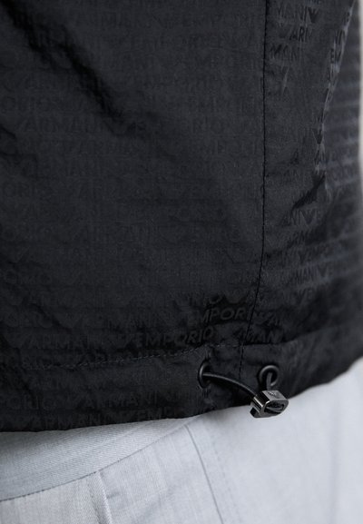 Casaco puffer preto com o logo "Emporio Armani" em relevo, apresentando uma bainha ajustável, textura suave e elementos de design elegantes.