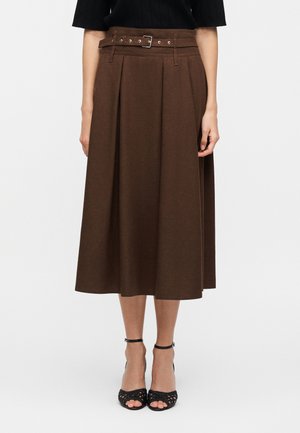 Femme portant une jupe midi marron taille haute à plis avec une ceinture assortie, associée à des talons noirs ouverts avec bride à la cheville.